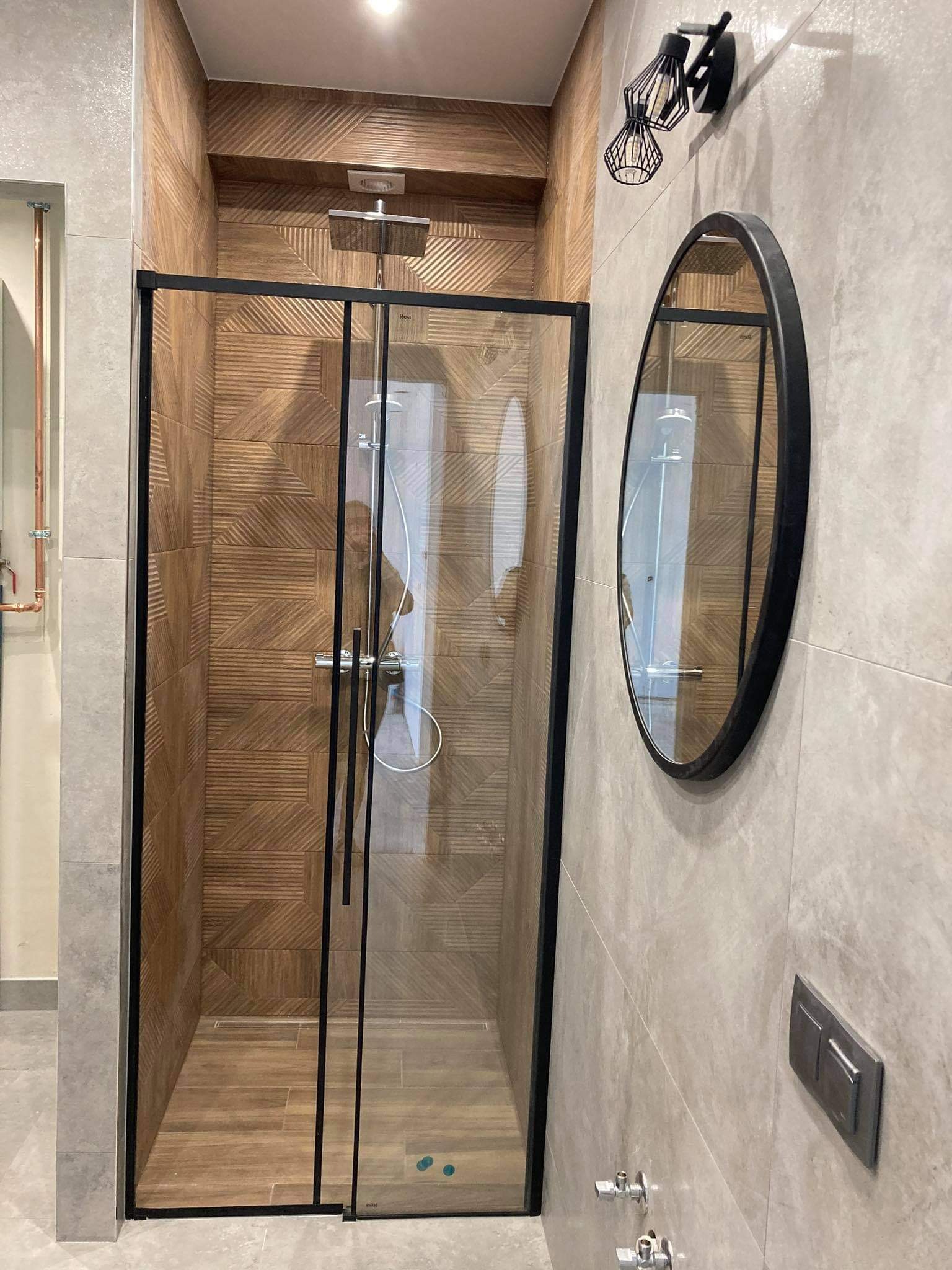 Nowoczesna kabina prysznicowa z czarną ramą, drewnopodobne płytki na ścianach, okrągłe lustro i designerska lampa na ścianie wykończonej szarymi płytkami.