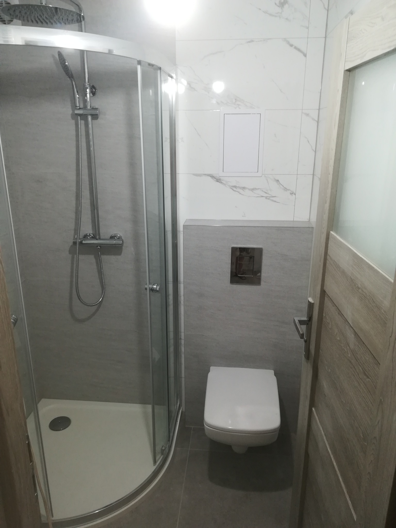 Nowoczesna łazienka z kabiną prysznicową z deszczownicą, podwieszaną toaletą i marmurowymi płytkami na ścianach, widok na fragment drzwi.