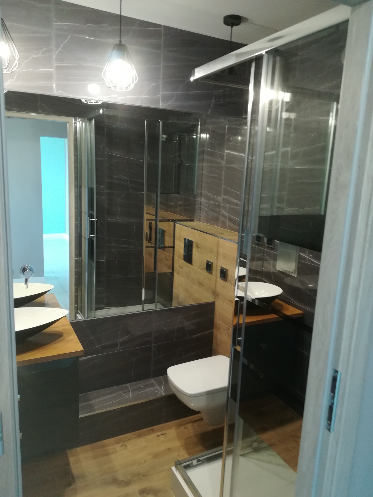 Nowoczesna łazienka z szarymi płytkami imitującymi marmur, drewnianymi akcentami i kabiną prysznicową ze szklanymi drzwiami przesuwnymi, widok zza uchylonych drzwi.