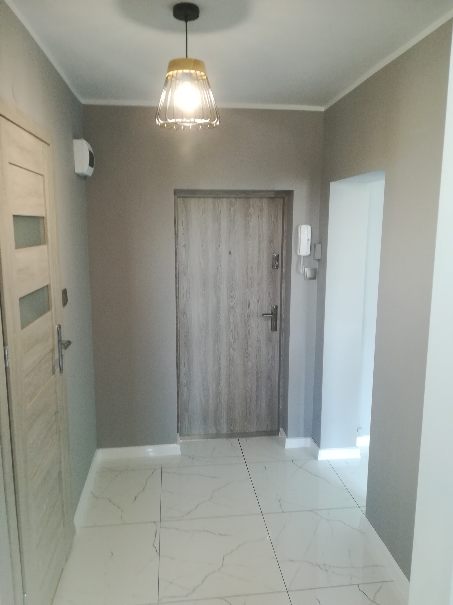 Jasnoszary przedpokój z białymi płytkami imitującymi marmur, lampą wiszącą z kloszem i dwiema parami drzwi w odcieniach szarości.