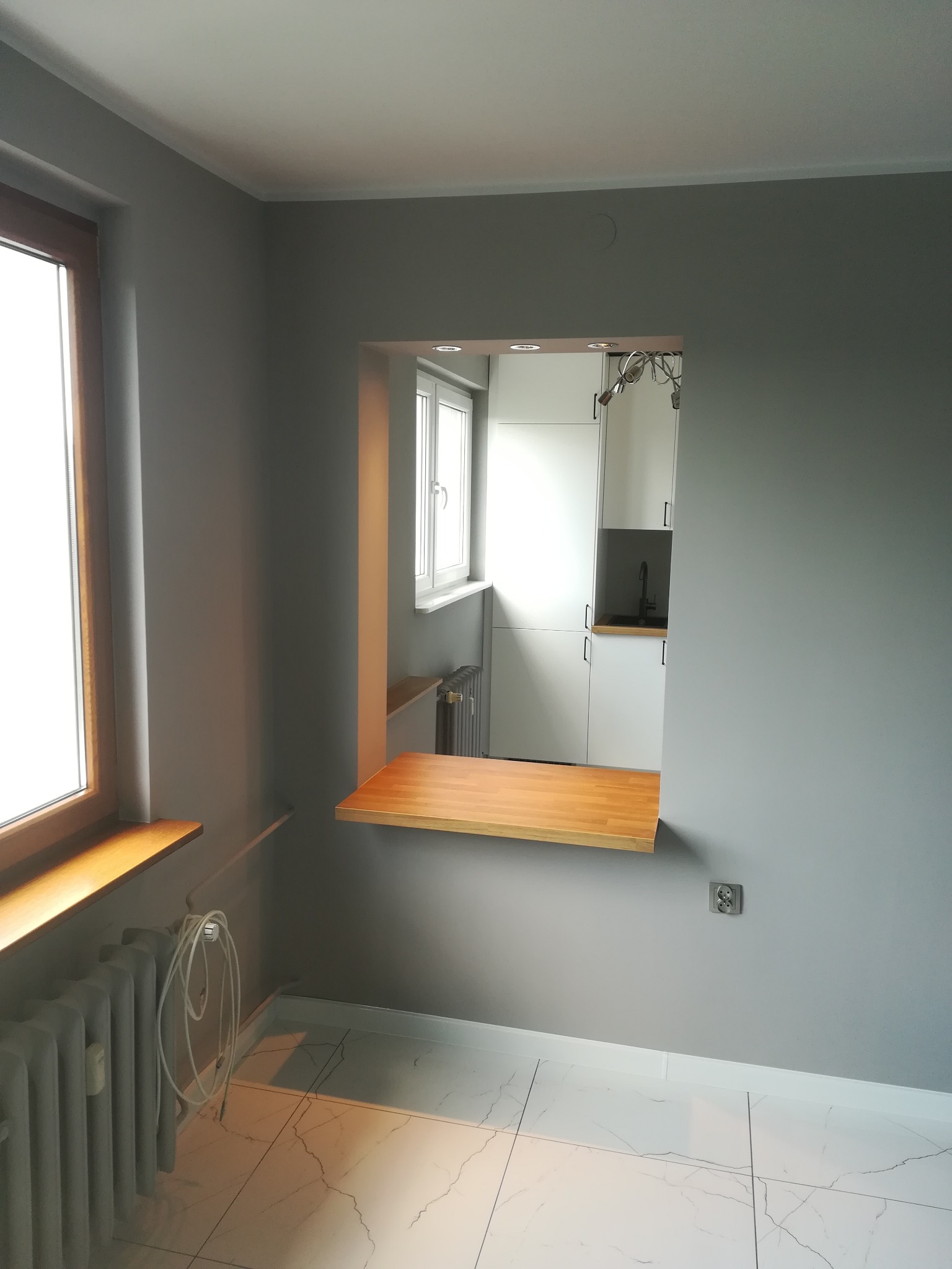 Szare wnętrze połączone z kuchnią przez okno z drewnianym blatem, widoczne białe szafki kuchenne i marmurowe płytki podłogowe.