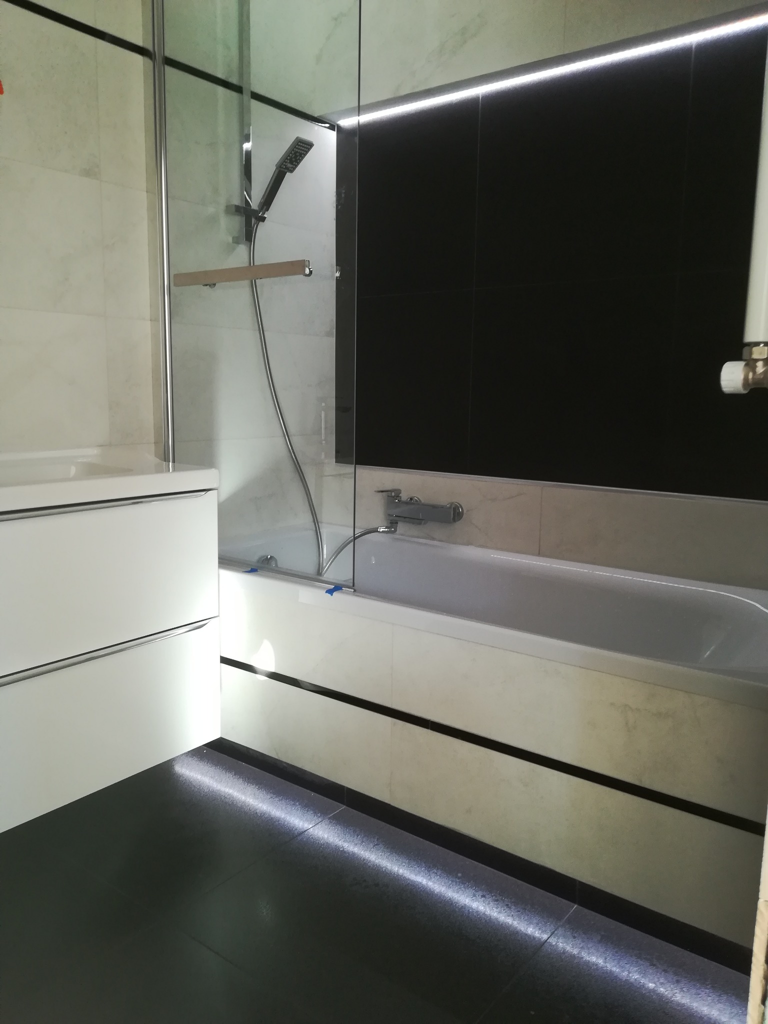 Nowoczesna łazienka z czarnymi płytkami, podświetlaną wanną, szklanym parawanem nawannowym i białą szafką umywalkową.