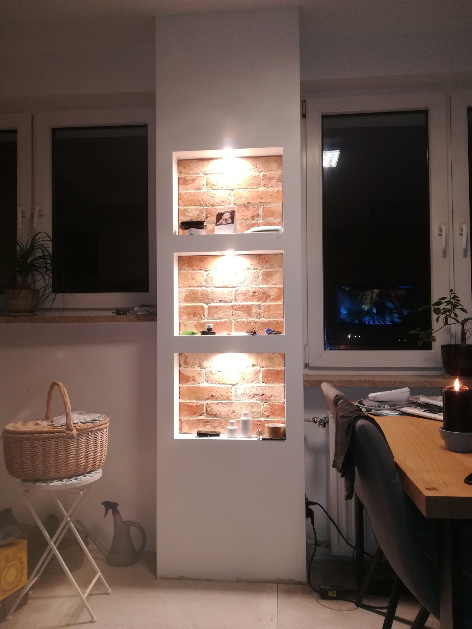 Wnęka ścienna z cegły, podświetlona trzema lampkami, z dekoracjami i przedmiotami na półkach, w tle okno z widokiem na nocne miasto.