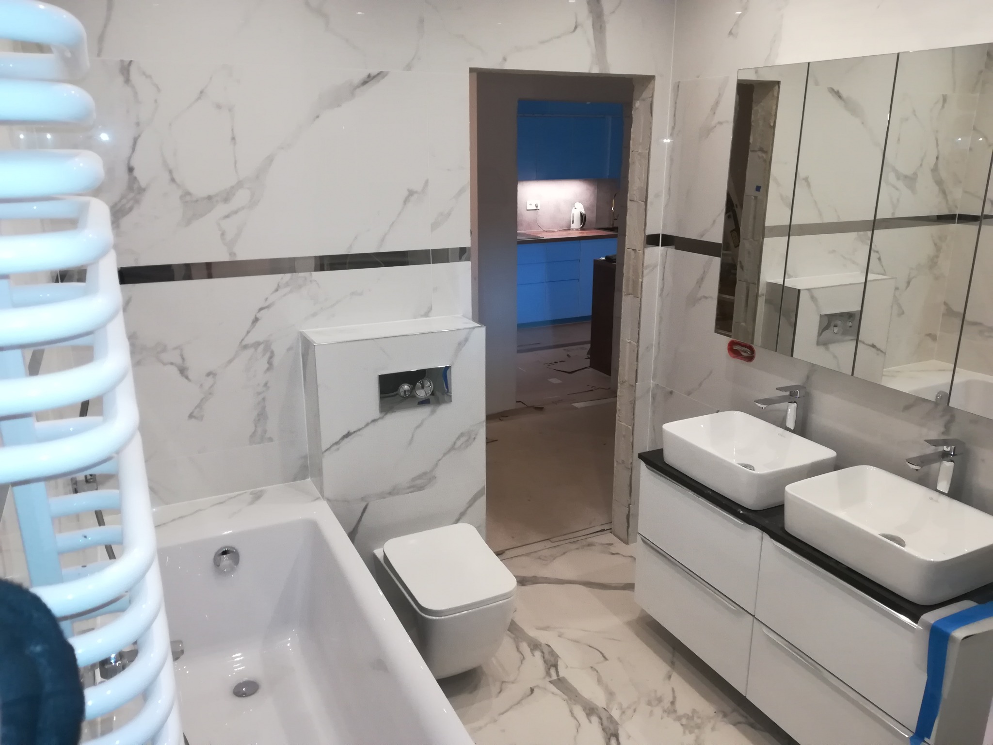 Nowoczesna łazienka z białymi płytkami imitującymi marmur, podwieszana toaleta, biała wanna, grzejnik drabinkowy i szafka z dwiema umywalkami nablatowymi, widok na nieukończone przejście do kuchni...