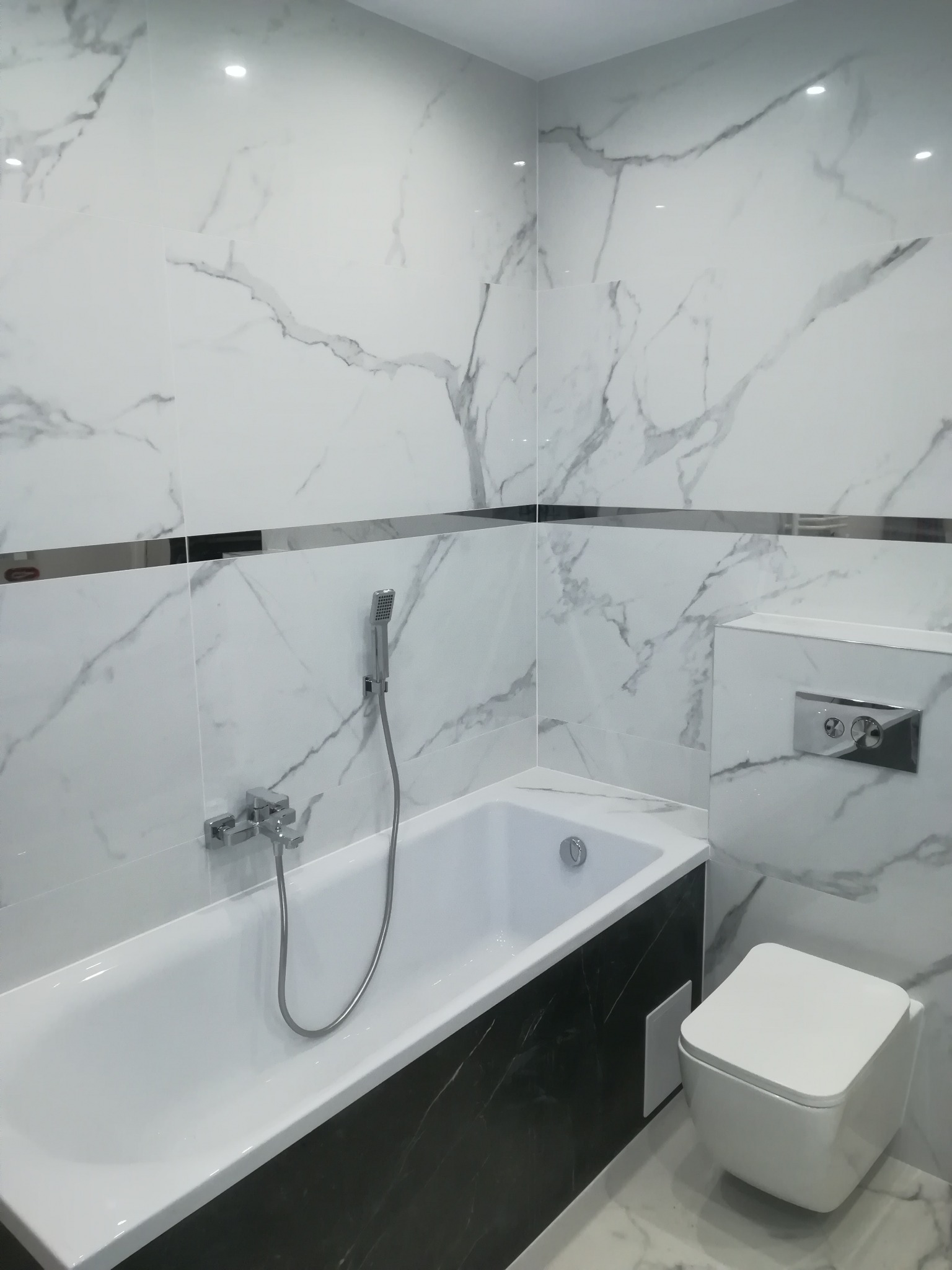 Nowoczesna łazienka z białą wanną obudowaną czarnym marmurem, białe płytki imitujące marmur na ścianach z poziomym paskiem dekoracyjnym, podwieszana toaleta.