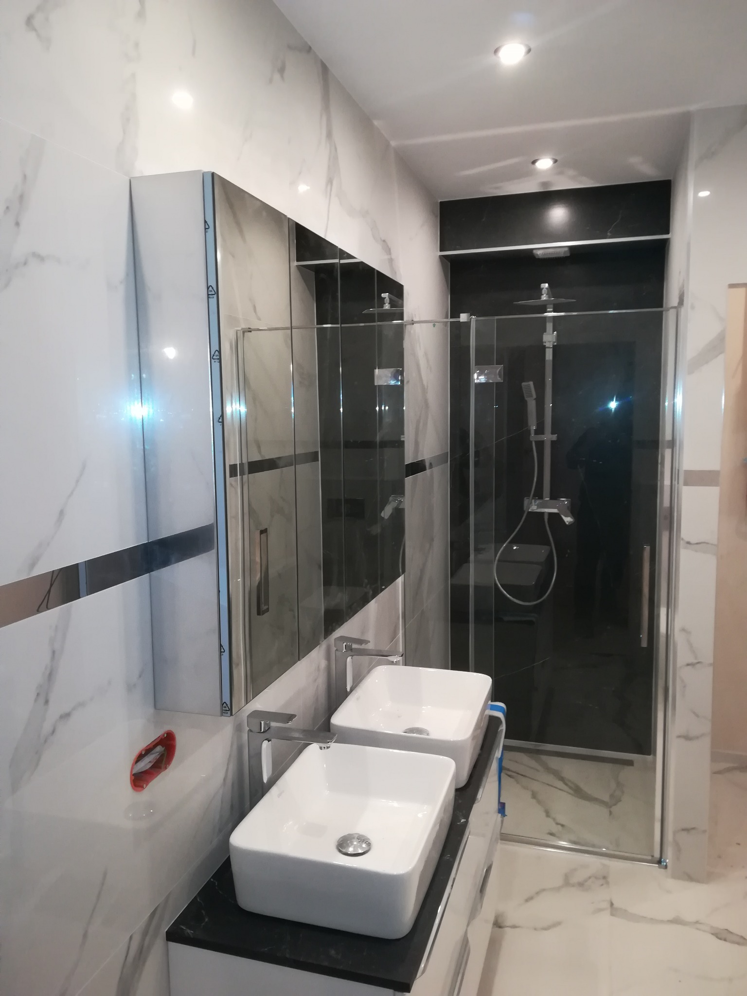 Nowoczesna łazienka z dwoma umywalkami nablatowymi, kabiną prysznicową typu walk-in z czarnym panelem, wykończona białymi płytkami imitującymi marmur z szarymi żyłami.