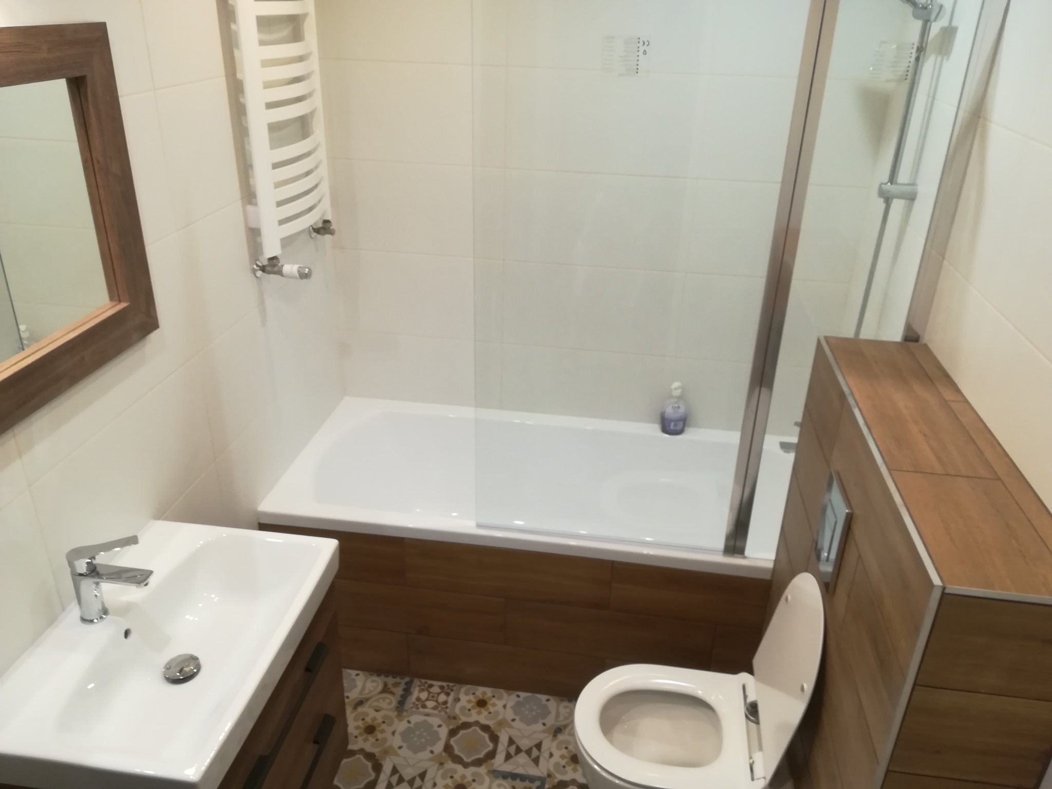 Nowoczesna łazienka po remoncie: biała wanna z parawanem, umywalka z szafką w odcieniach brązu, wisząca toaleta i dekoracyjna podłoga z wzorem geometrycznym.