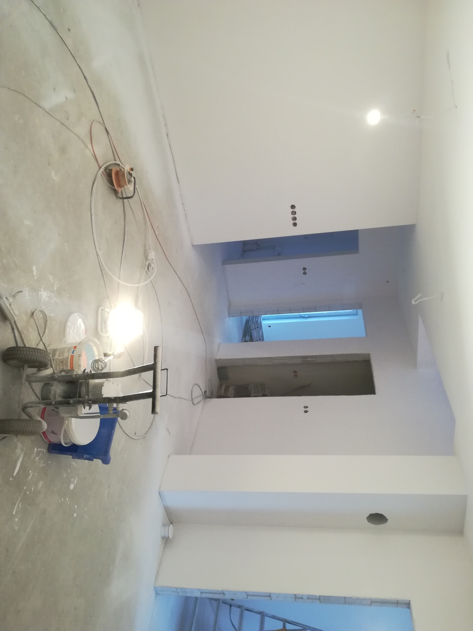 Wnętrze w trakcie malowania natryskowego, widoczny agregat malarski z niebieskim pojemnikiem, świeżo pomalowane na biało ściany i sufit, oraz przygotowane otwory na gniazdka i oświetlenie.