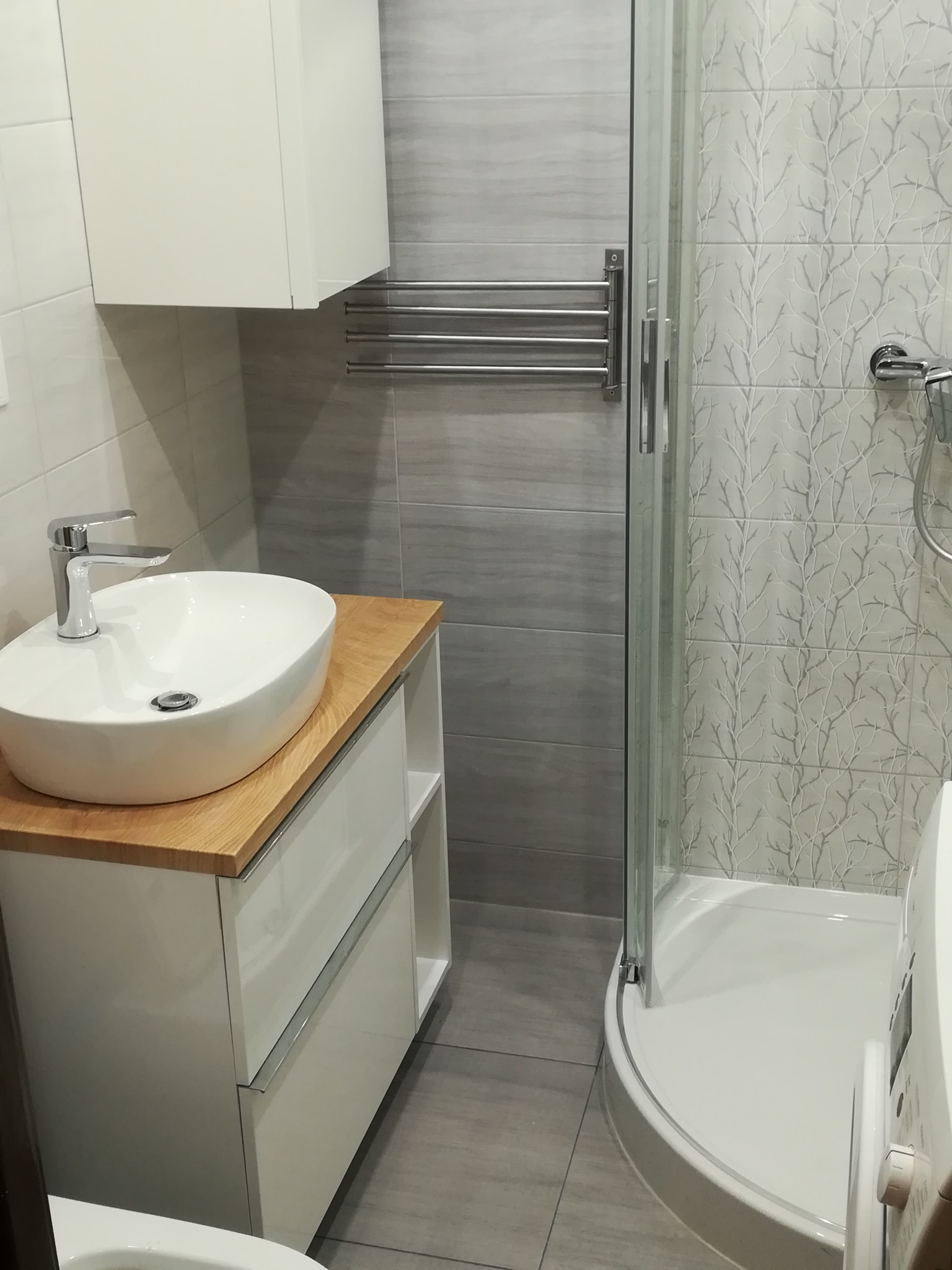 Nowoczesna łazienka z umywalką nablatową na drewnianym blacie, kabiną prysznicową z wzorzystymi płytkami i grzejnikiem drabinkowym.