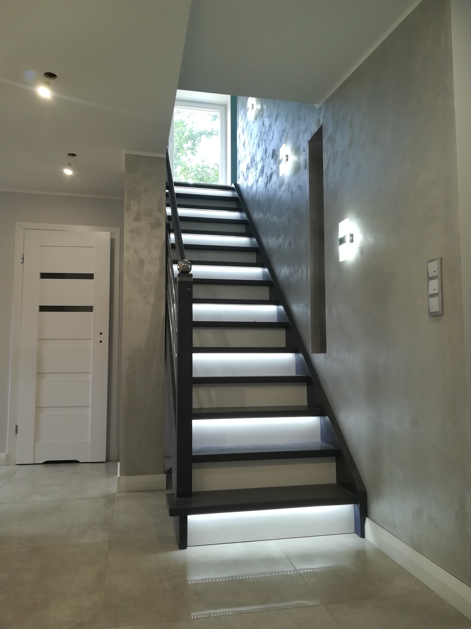 Nowoczesne schody z podświetleniem LED wzdłuż stopni, szara ściana z efektem dekoracyjnym, białe drzwi z czarnymi wstawkami.