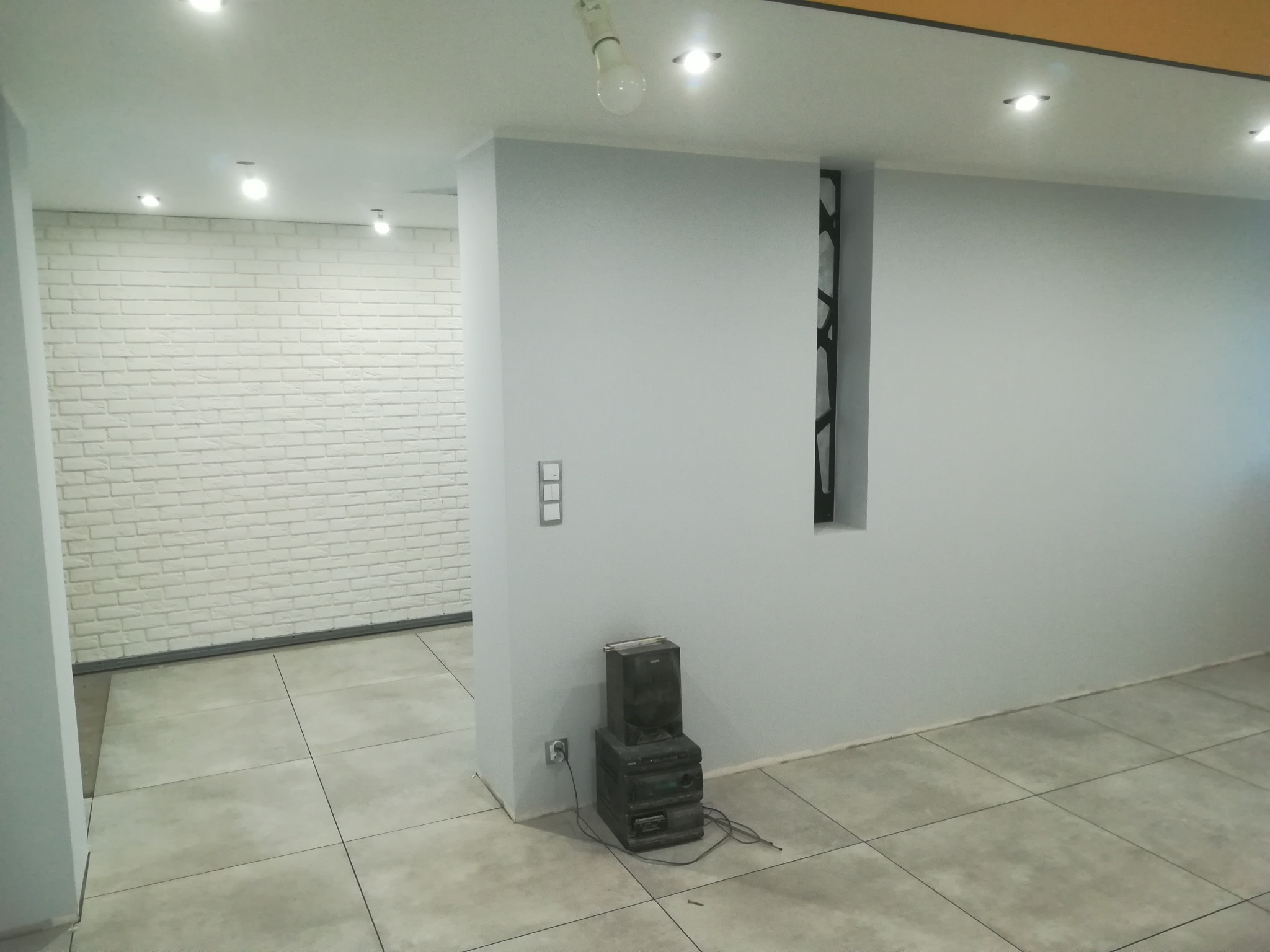 Wnętrze biura po odświeżeniu: ściany pomalowane na szaro, biała cegła dekoracyjna, szare płytki podłogowe, widoczny fragment instalacji elektrycznej i starego sprzętu audio.