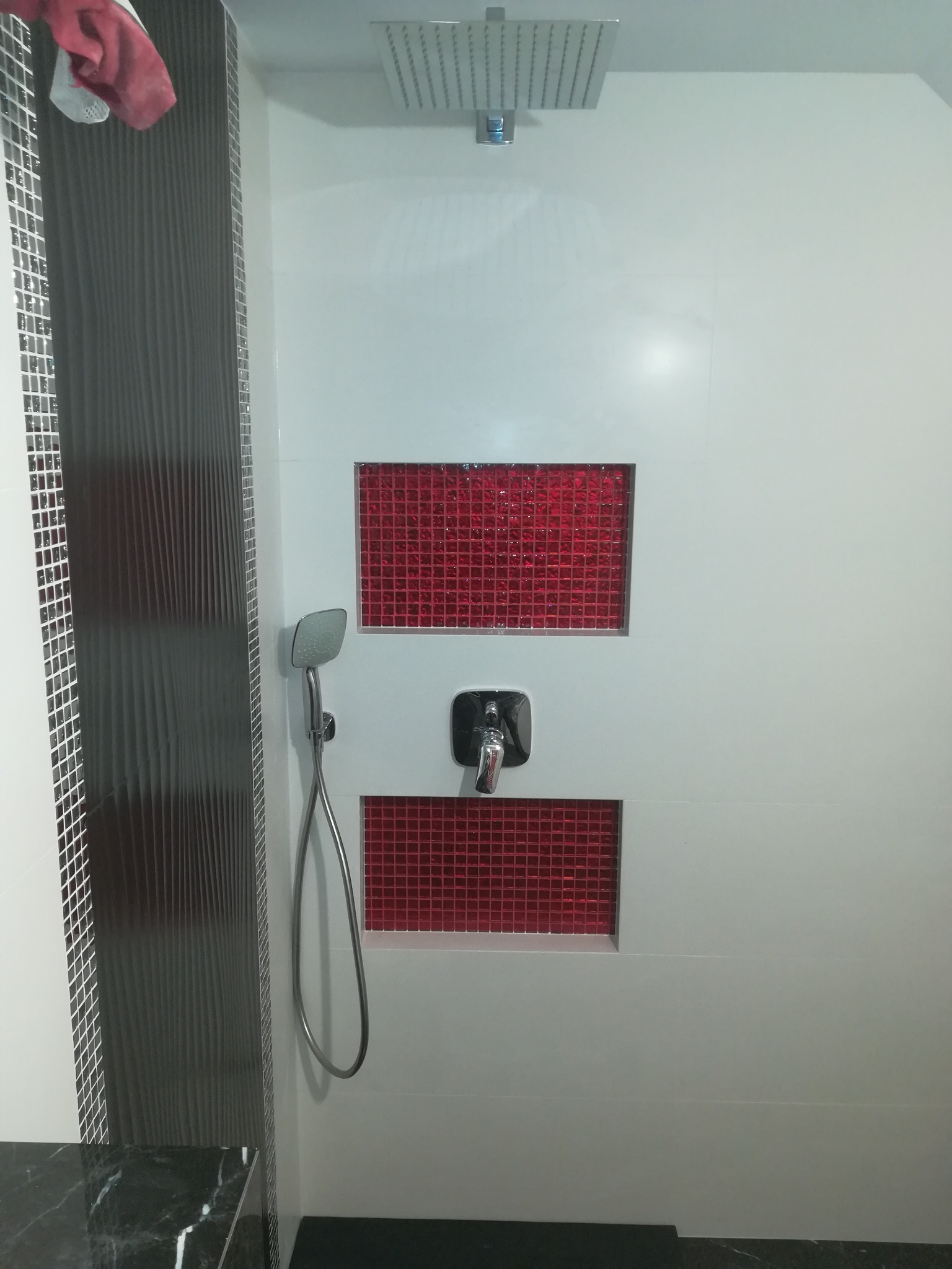 Nowoczesna kabina prysznicowa z białymi ścianami, wstawkami z czerwonej mozaiki, deszczownicą i baterią natryskową. Po lewej stronie widoczny fragment ściany z ciemnoszarą, pionowo żłobkowaną...