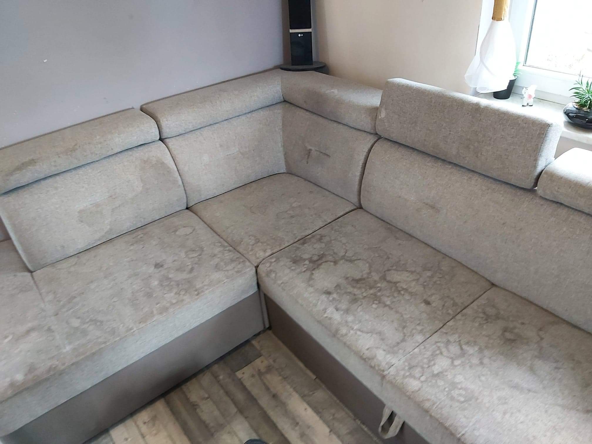 Zabrudzona, narożna kanapa w odcieniach szarości, wymagająca profesjonalnego czyszczenia tapicerki meblowej w Środzie Wielkopolskiej. Widoczne plamy i zacieki na siedzisku i oparciach.