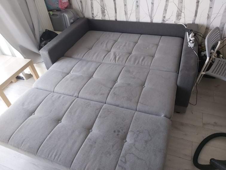 Rozłożona szara kanapa z widocznymi plamami na tapicerce, na podłodze leży wąż od odkurzacza.