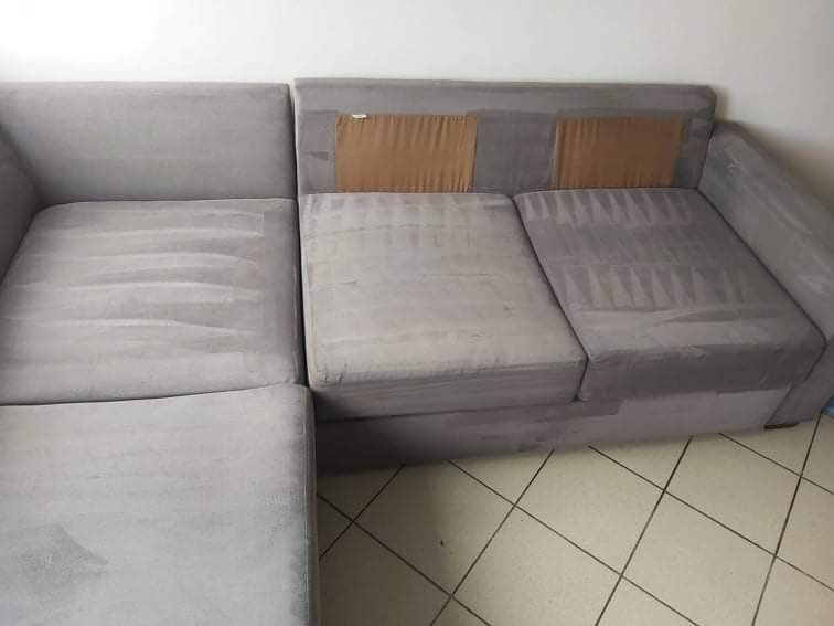 Szara, narożna kanapa z widocznymi śladami po czyszczeniu tapicerki, z brązowymi zabezpieczeniami na oparciu.