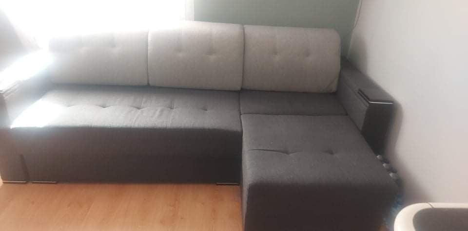 Szaro-beżowa, rozkładana kanapa narożna z widocznymi guzikami na siedzisku, ustawiona na drewnianej podłodze, z jasnymi zagłówkami.