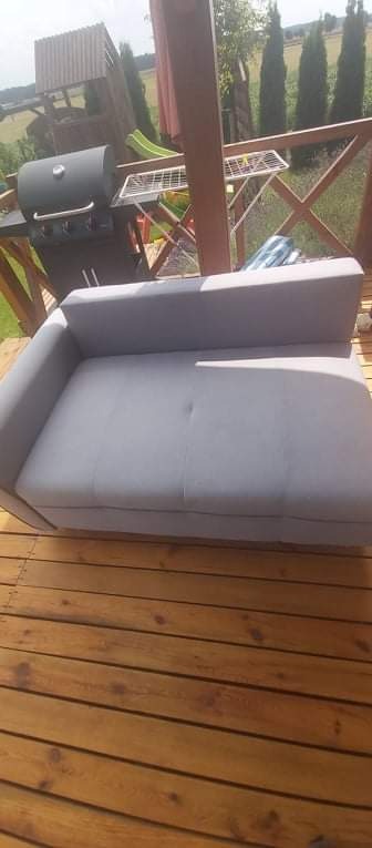 Szara, dwuosobowa kanapa na drewnianym tarasie z grillem i suszarką do ubrań w tle.