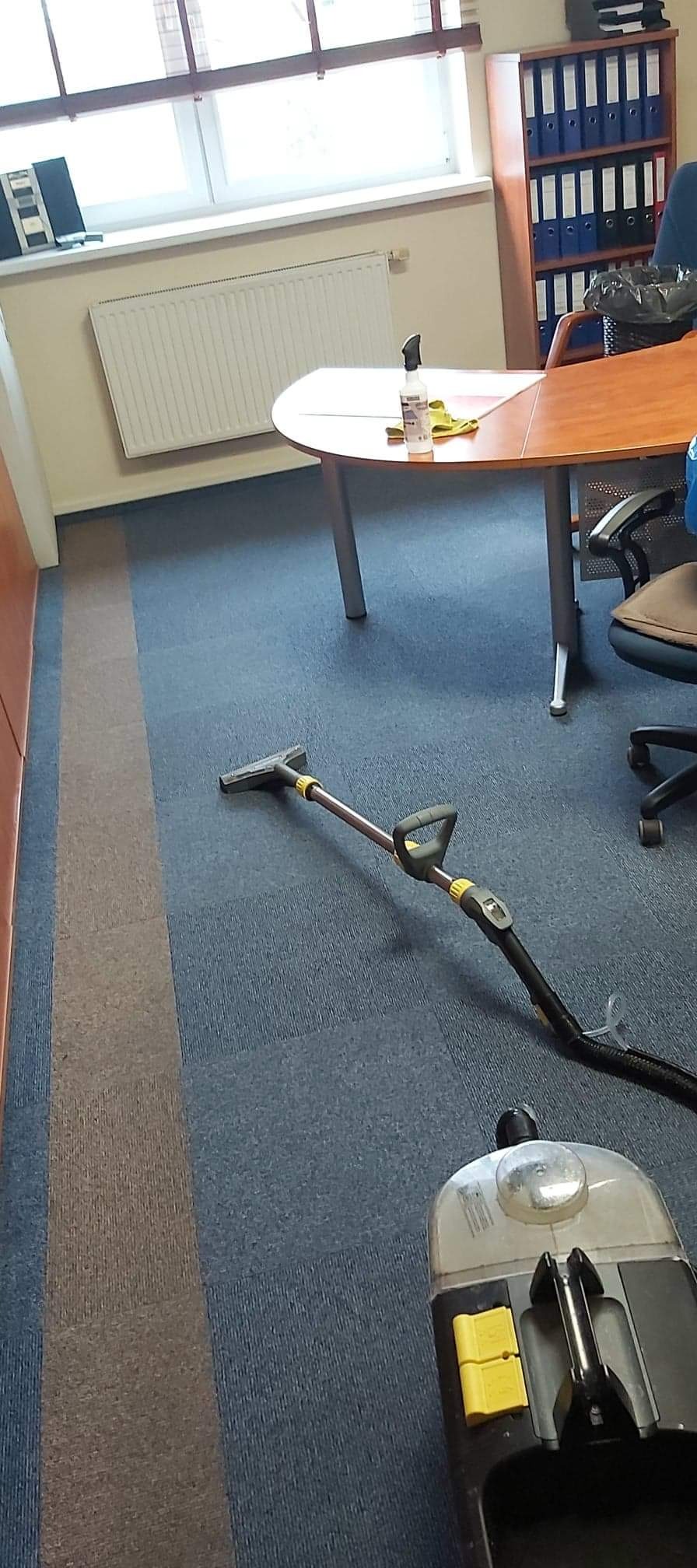 Profesjonalny odkurzacz piorący na niebiesko-szarym dywanie w biurze, częściowo wyczyszczony fragment dywanu z widoczną różnicą koloru, biurko i regał z segregatorami w tle.