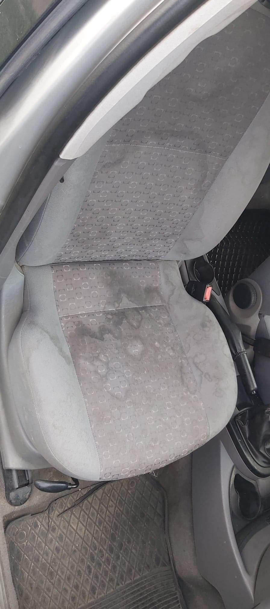 Szare, tekstylne siedzenie samochodowe z widocznymi plamami przed czyszczeniem, obok czarna gumowa wycieraczka samochodowa.