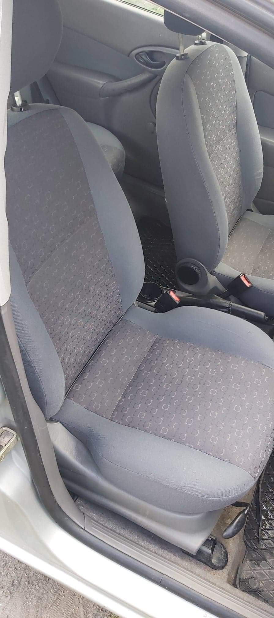 Wnętrze samochodu: szare fotele z wzorem w małe kwadraty, widoczne pasy bezpieczeństwa i gumowa wycieraczka pod nogami kierowcy.
