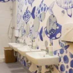 Rząd czterech białych umywalek z chromowanymi kranami zamontowanych na ścianie z artystycznym motywem niebieskich ryb w łazience publicznej z białymi panelami ściennymi i metalowym koszem na śmieci.