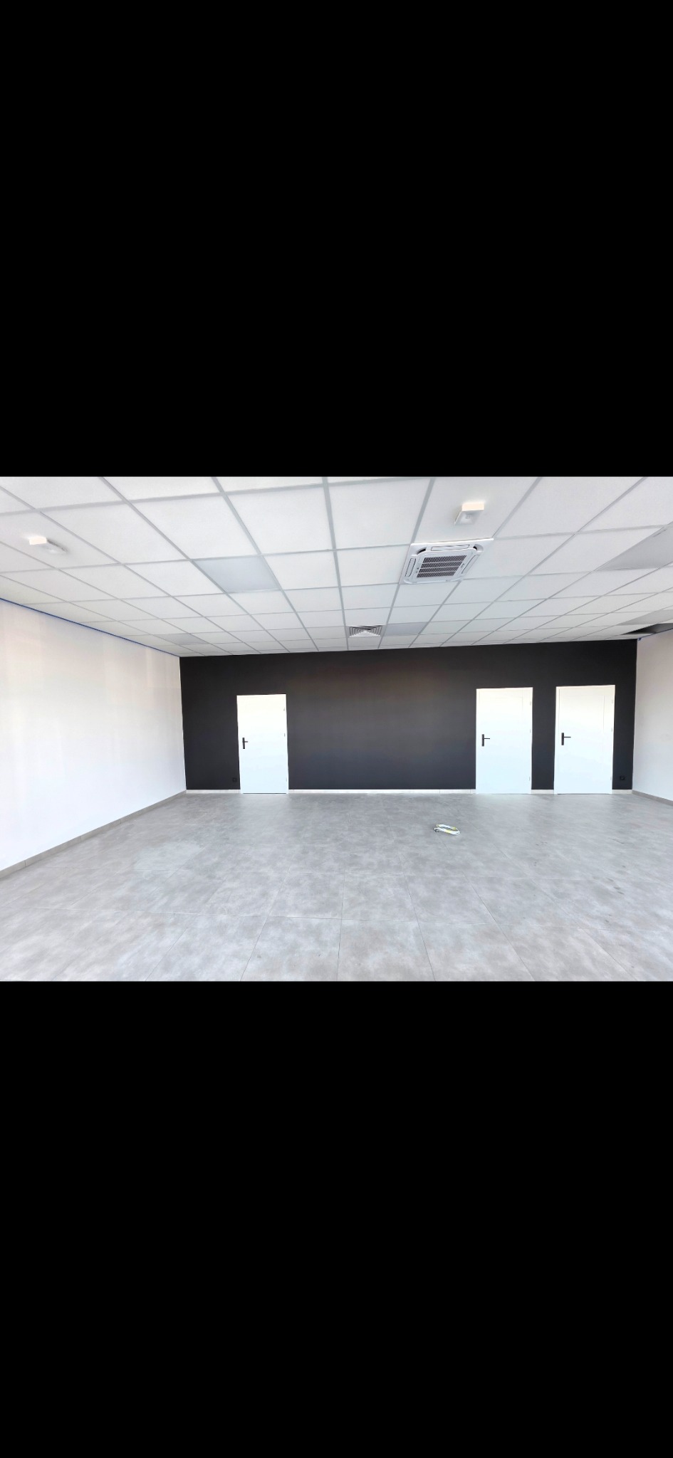 Wnętrze biura po remoncie: szara podłoga, czarna ściana z trzema białymi drzwiami, białe ściany boczne i sufit kasetonowy z klimatyzacją. Minimalistyczny design.