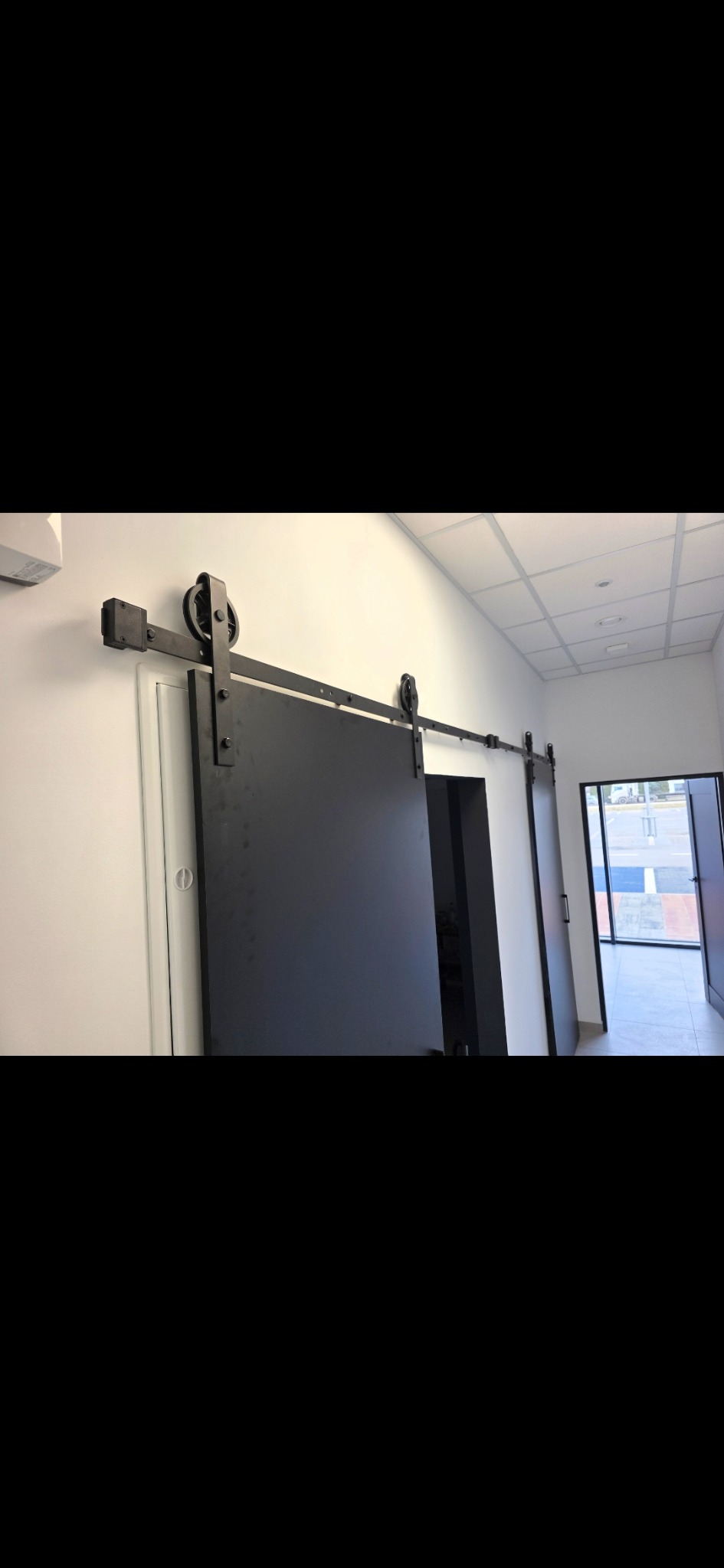 Nowoczesne, przesuwne drzwi w stylu loftowym, zamontowane na metalowej szynie z widocznymi rolkami. Minimalistyczne wnętrze biura z białymi ścianami i sufitową kratownicą.