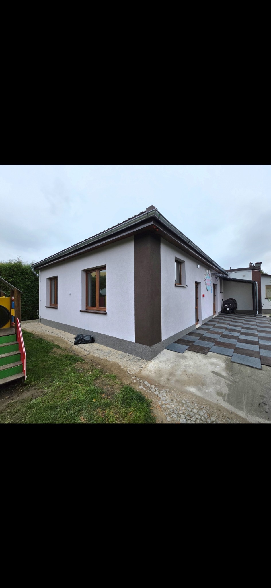 Ocieplony budynek z brązowym akcentem na elewacji, widoczny fragment placu zabaw i kostka brukowa. Ujęcie z poziomu gruntu, pochmurne niebo.