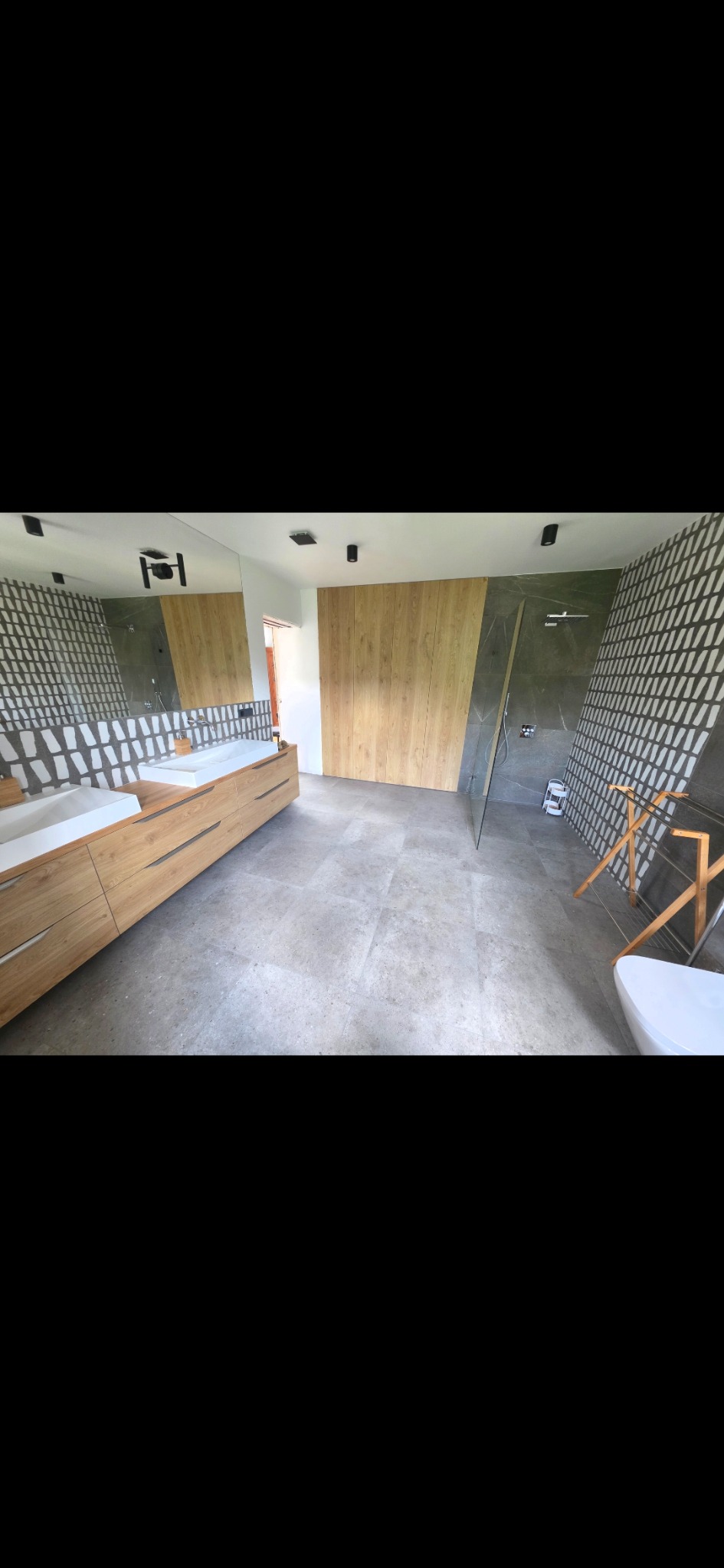 Nowoczesna łazienka z drewnianą zabudową umywalki, szarą podłogą i geometrycznymi wzorami na ścianach. Kabina prysznicowa ze szklaną ścianką. Minimalistyczny design.