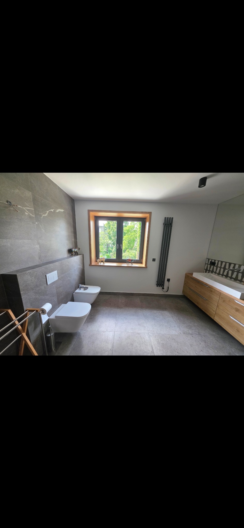 Nowoczesna łazienka z szarymi płytkami, drewnianą szafką pod umywalką i oknem z widokiem na zieleń. Minimalistyczny design z wiszącą toaletą i bidetem.