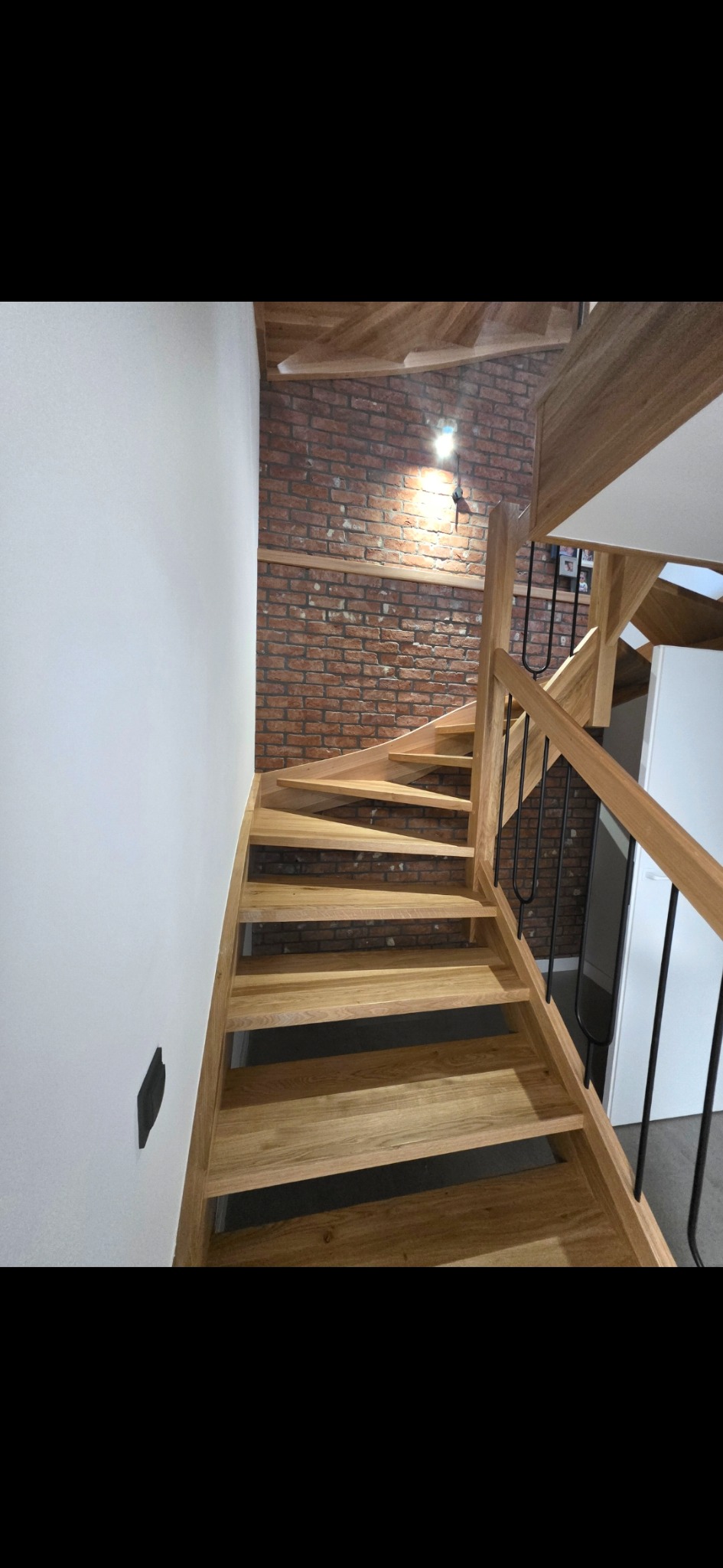 Drewniane schody spiralne z czarną balustradą, ściana z cegły w tle, oświetlenie punktowe. Widok z dołu, nowoczesny design.