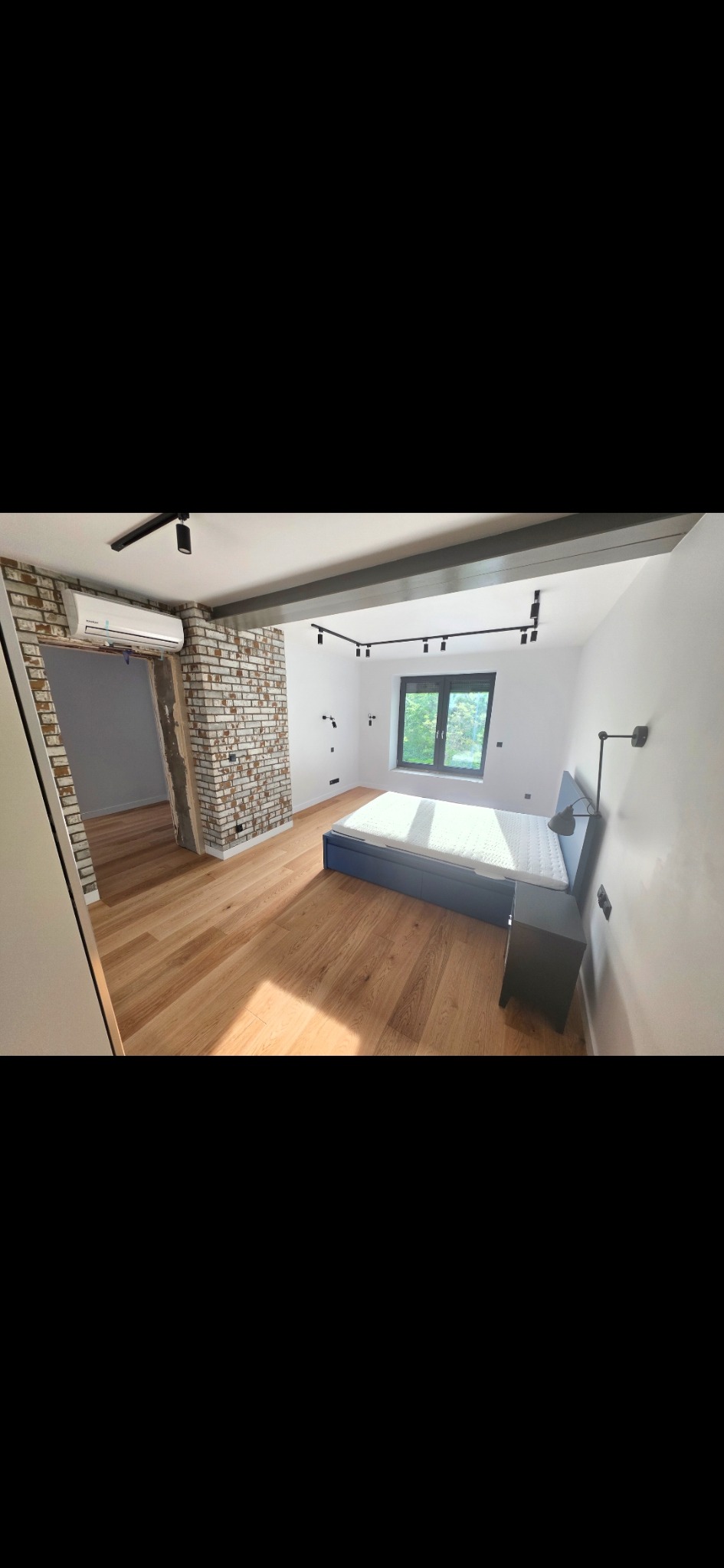 Jasne wnętrze sypialni po remoncie: drewniana podłoga, ceglana ściana z klimatyzacją, czarne oświetlenie szynowe i okno. Widoczny fragment łóżka i szafki nocnej.