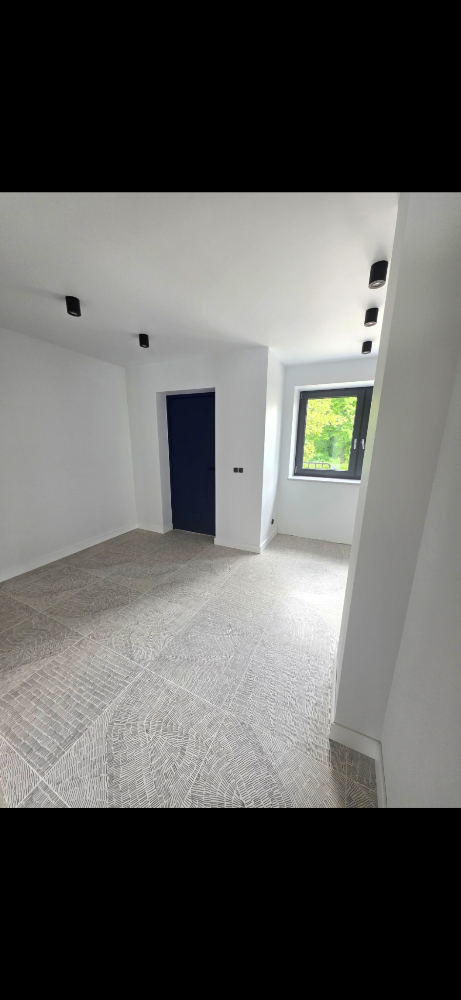 Wykończone wnętrze: wzorzysta podłoga z płytek, białe ściany, czarne lampy sufitowe i ciemne drzwi. Widok na zieleń za oknem. Minimalistyczny design.