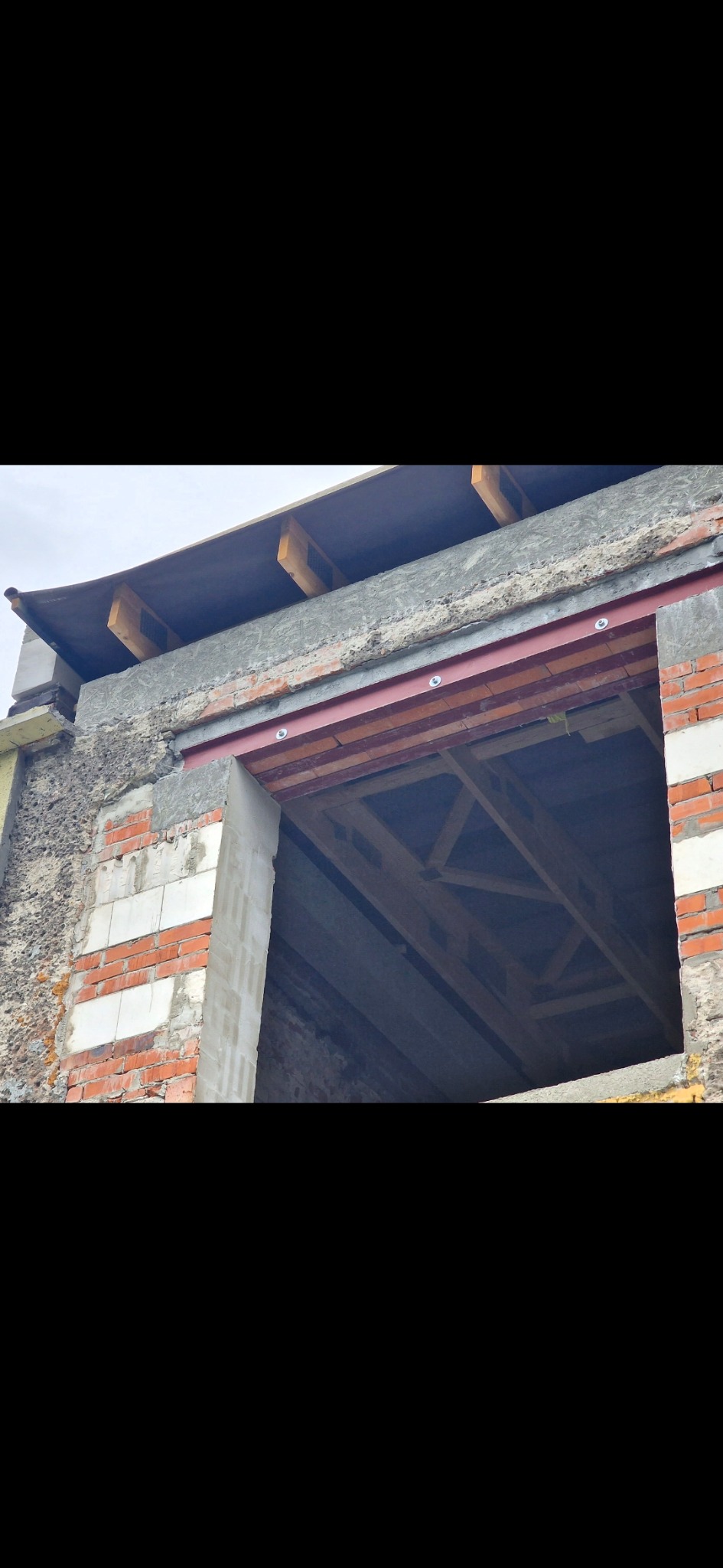 Surowy stan otworu okiennego w murze z cegły i betonu, z widoczną konstrukcją drewnianą wewnątrz. Elementy stalowe wzmacniające nadproże. Prace murarskie w toku.