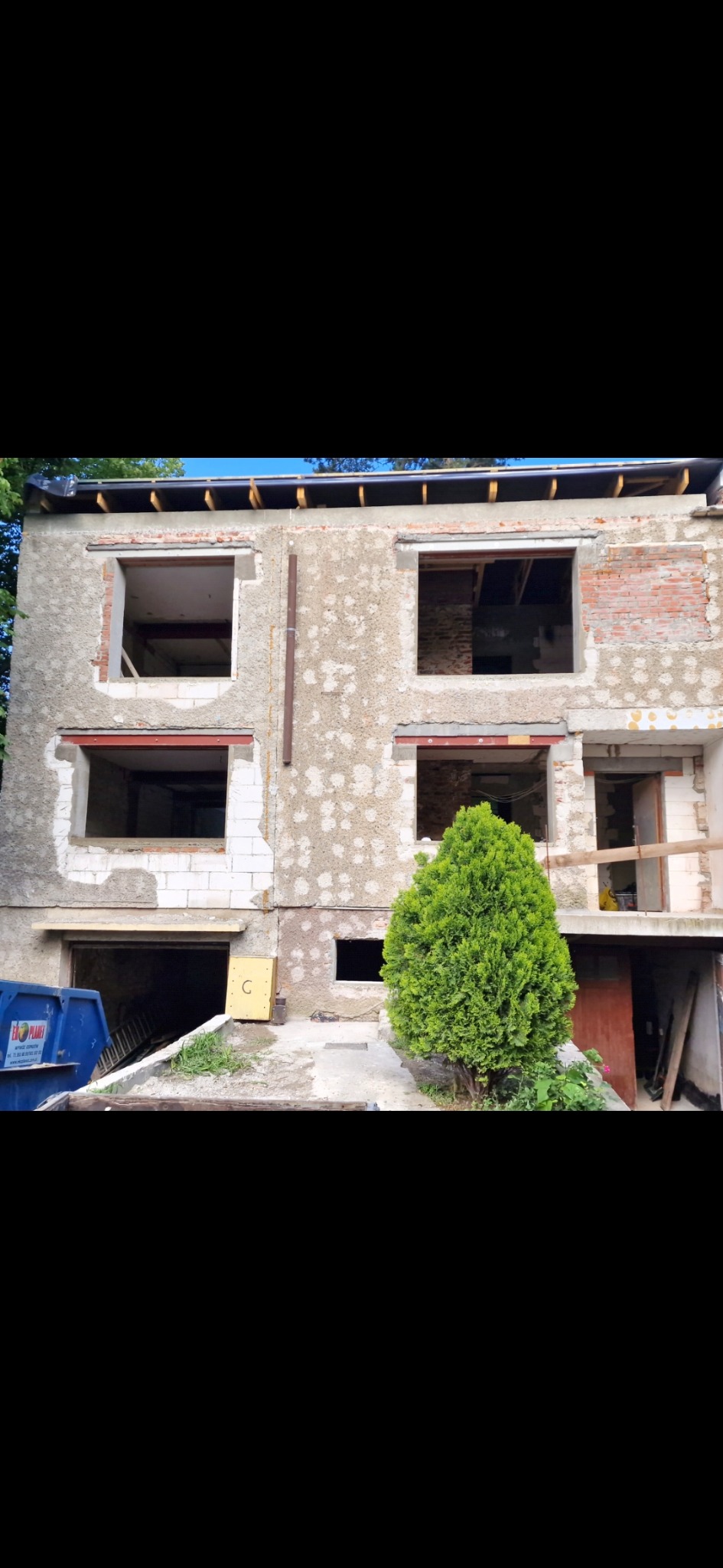 Dwupiętrowy budynek w trakcie remontu, z odsłoniętą cegłą i pustymi otworami okiennymi. Na pierwszym planie zielony krzew i kontener na gruz.