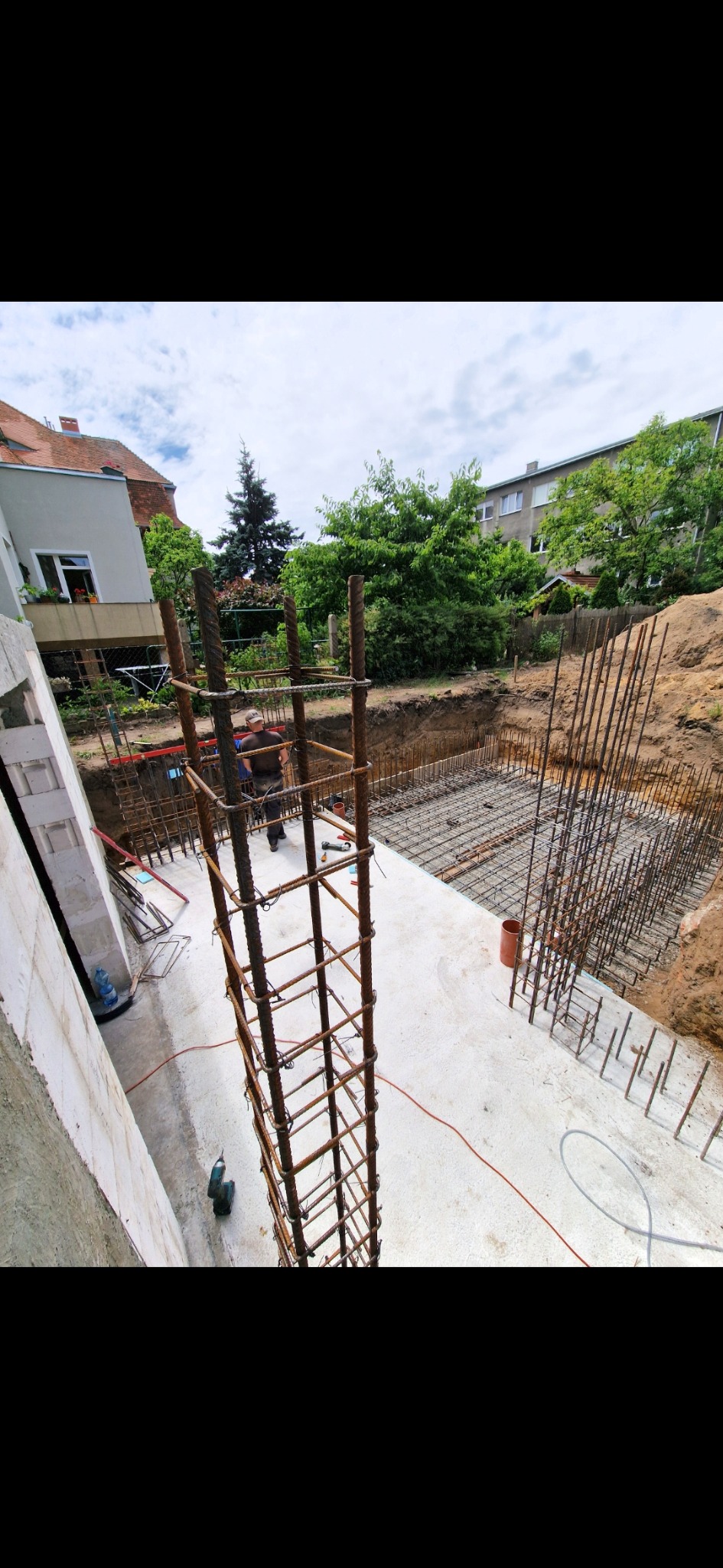 Zbrojenie fundamentów pod budynek jednorodzinny. Widoczne stalowe pręty, siatka zbrojeniowa i wykop. W tle budynki mieszkalne i zieleń. Prace budowlane w toku.