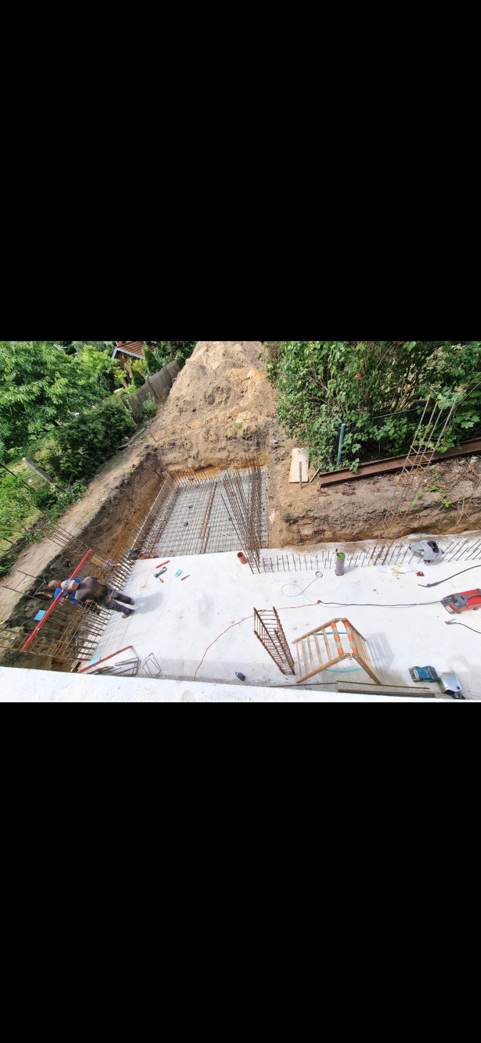 Z góry widok na wykopany dół z betonową płytą fundamentową i zbrojeniem. Widoczni pracownicy i narzędzia budowlane. Tło: ziemia i zieleń.