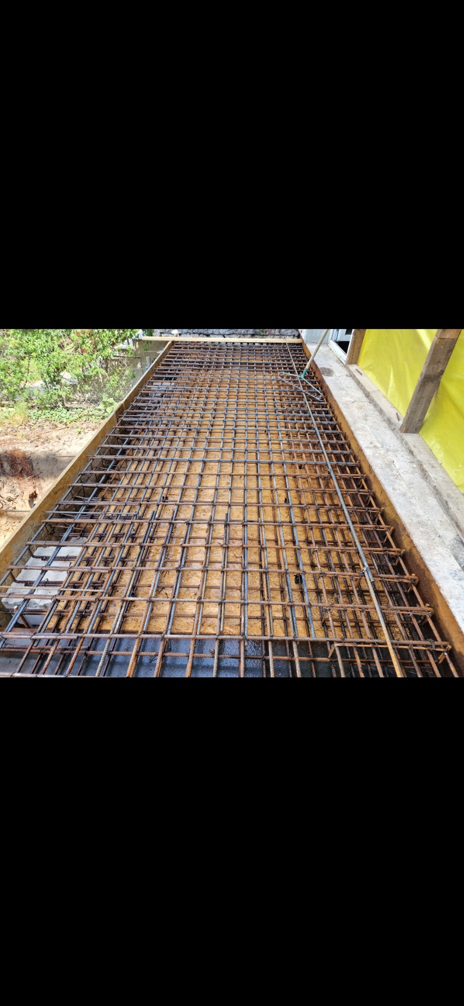 Zbrojenie fundamentu z siatki stalowej w drewnianym szalunku, przygotowane do zalania betonem. Widoczne elementy konstrukcyjne i otoczenie budowy.