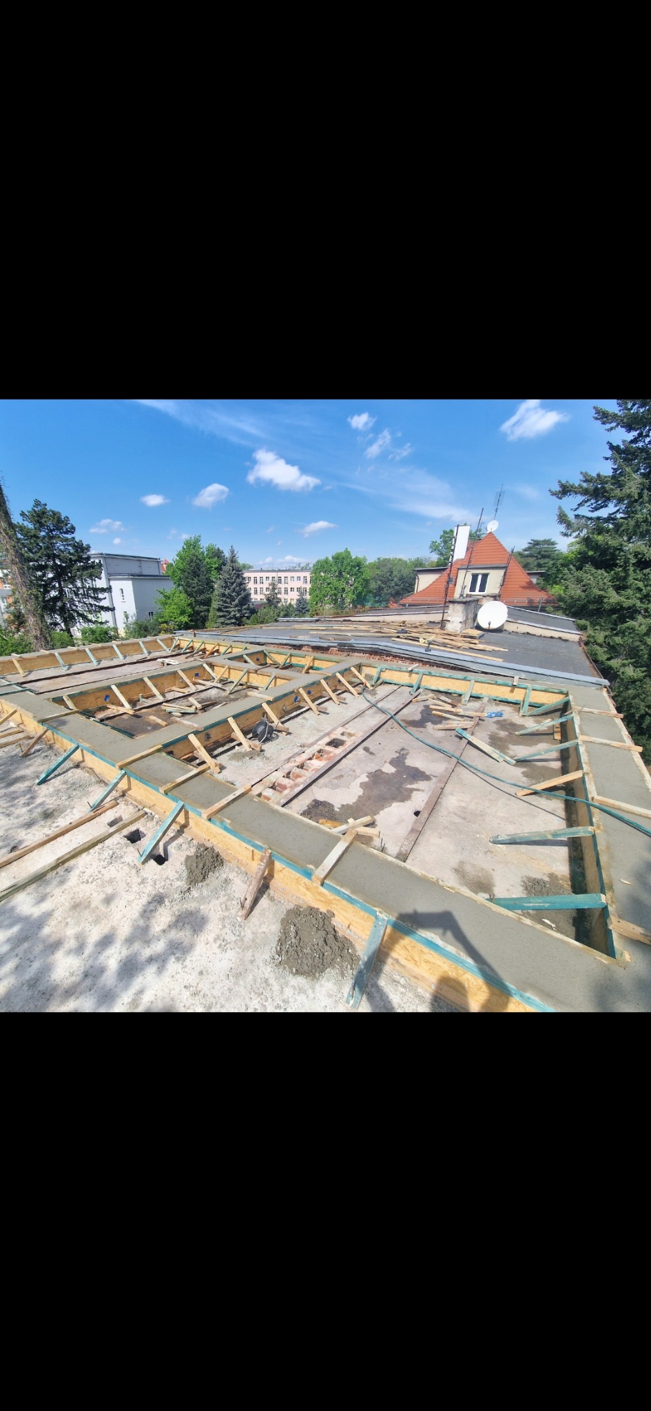 Zalewanie betonem szalunków na dachu budynku w słoneczny dzień. Widoczne drewniane elementy konstrukcyjne i świeży beton. W tle budynki i drzewa.