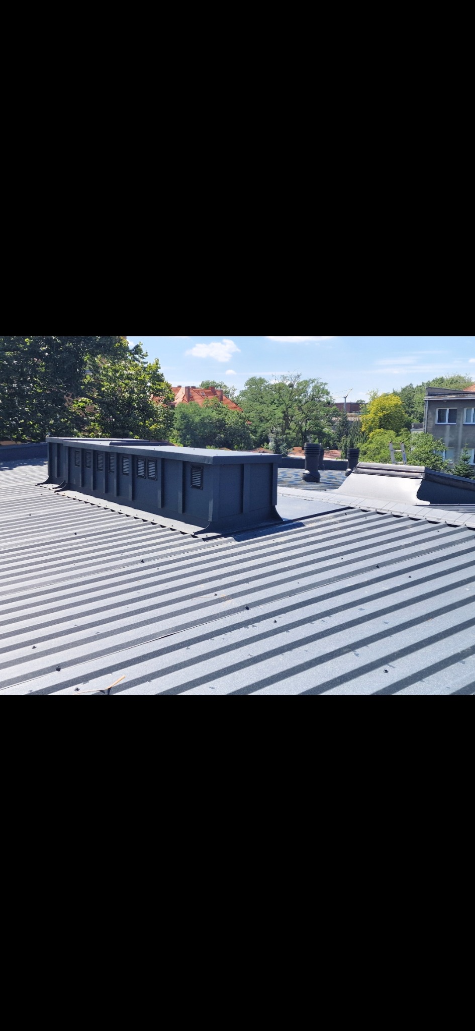 Szary dach z blachy trapezowej z elementami wentylacyjnymi na tle zieleni drzew i budynków w słoneczny dzień. Widok z góry.