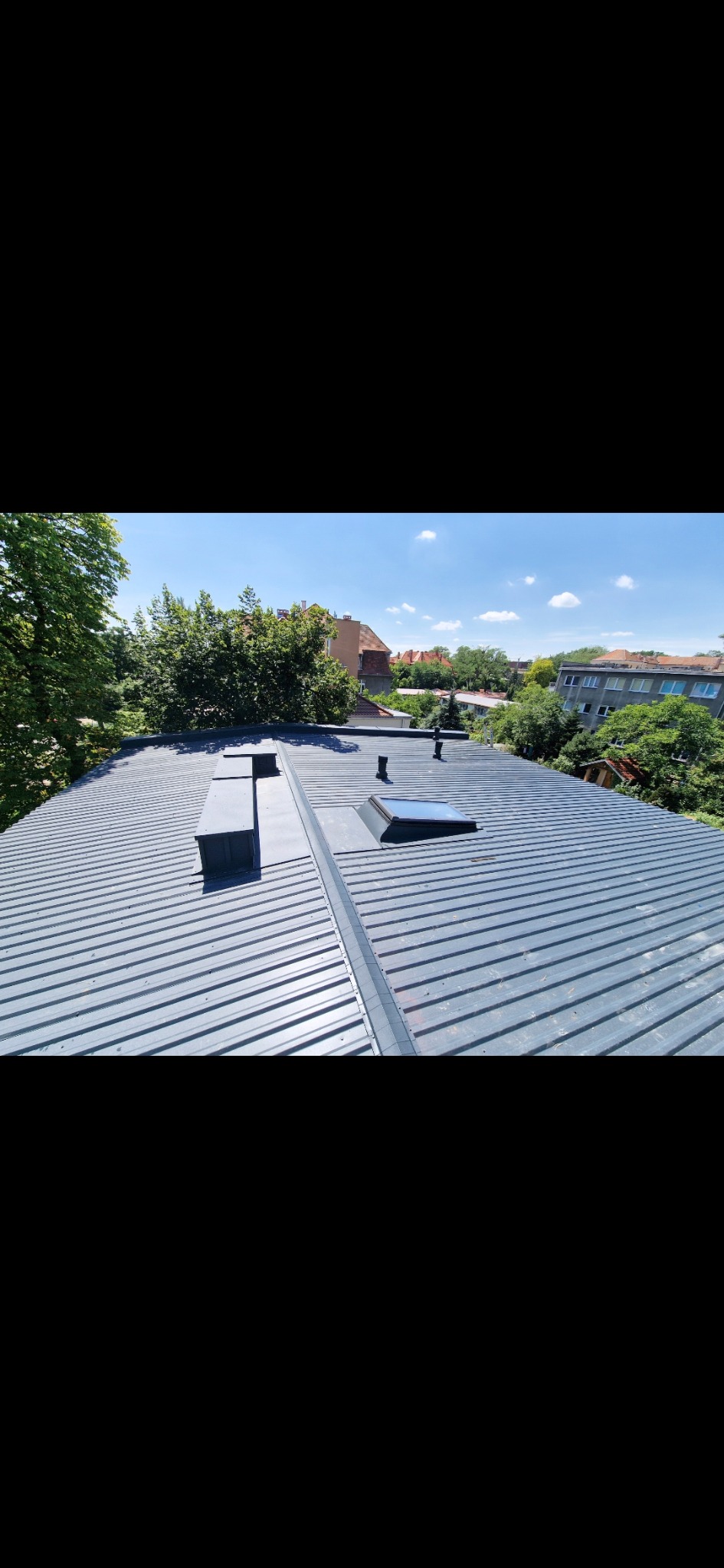 Szary blaszany dach z kominem i oknem dachowym na tle zieleni i zabudowań miejskich w słoneczny dzień. Widok z góry.