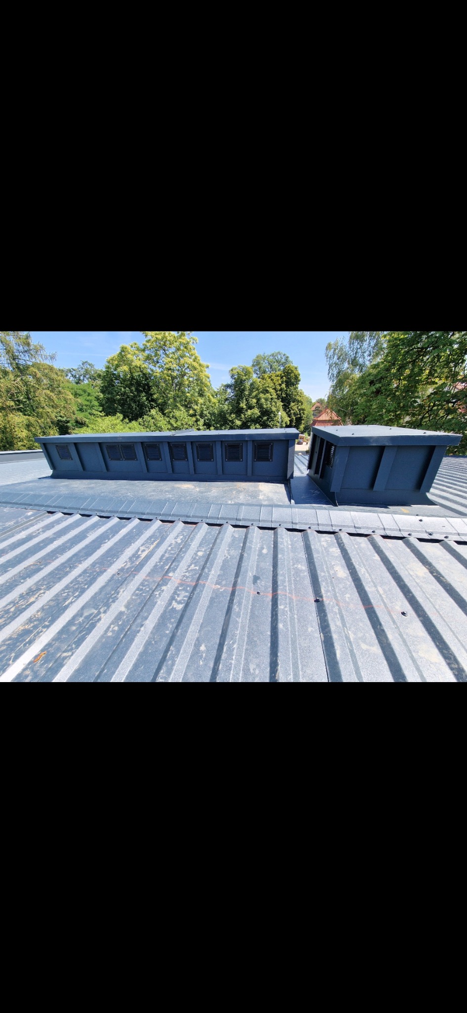 Metalowy dach z dwoma świetlikami dachowymi w kolorze grafitowym, widok z góry, w tle zieleń drzew i błękitne niebo. Dach pokryty blachą trapezową.