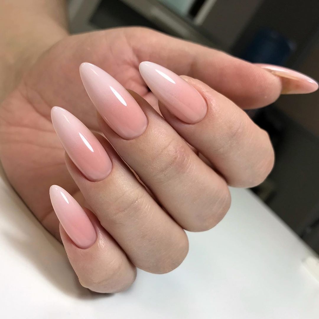 Dłoń z długimi, migdałowymi paznokciami pomalowanymi lakierem hybrydowym w odcieniach różu i bieli, tworzącym efekt ombre.