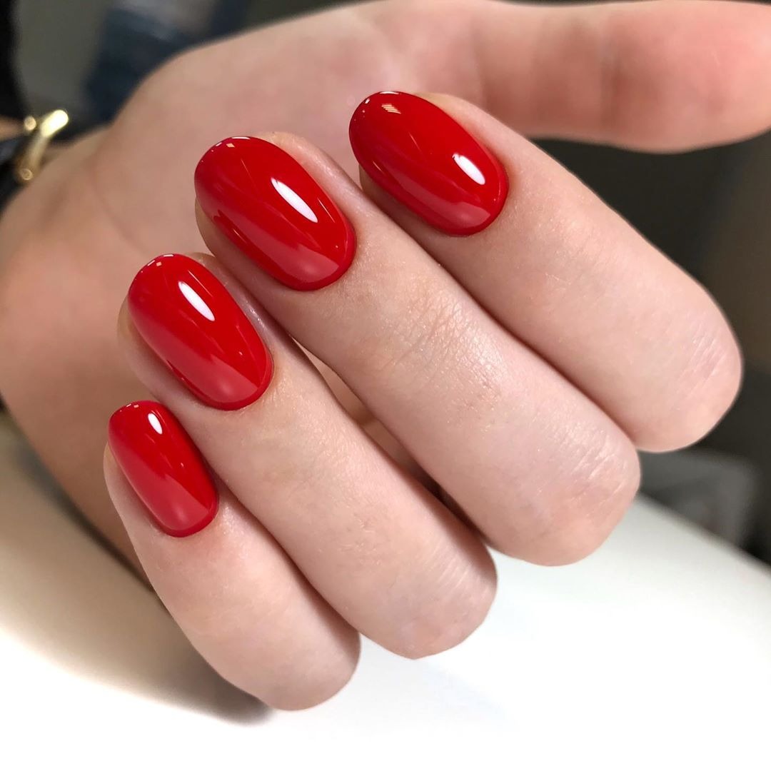 Dłoń z idealnie pomalowanymi, owalnymi paznokciami w intensywnym, błyszczącym odcieniu czerwieni. Widoczne delikatne refleksy światła na lakierze.