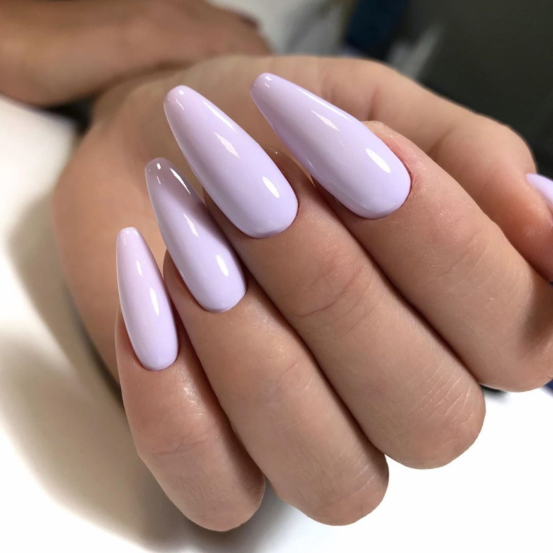 Dłoń z długimi, migdałowymi paznokciami pomalowanymi na pastelowy, liliowy kolor z wyraźnym połyskiem.