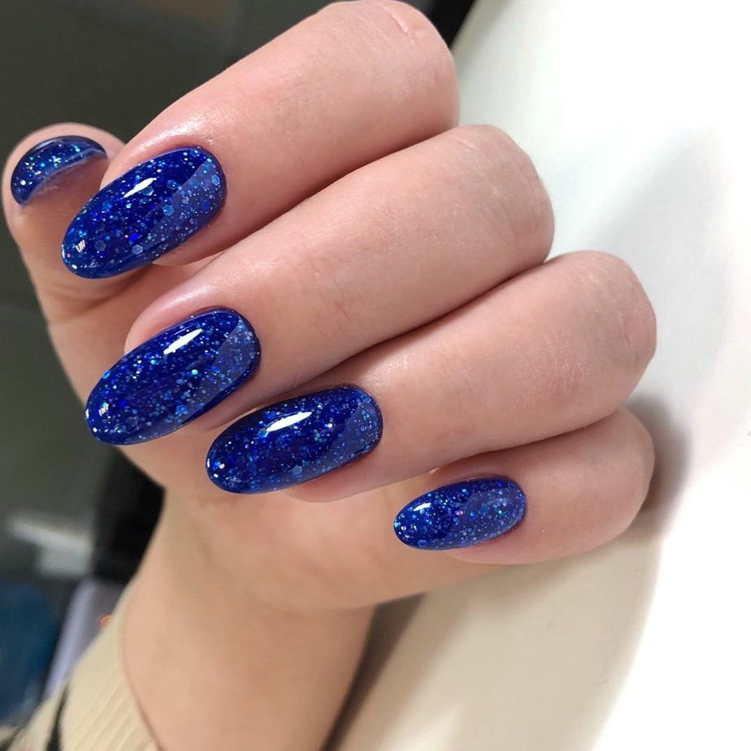 Dłoń z owalnymi paznokciami pokrytymi ciemnoniebieskim lakierem z brokatem, widoczne drobne niedoskonałości skórki.