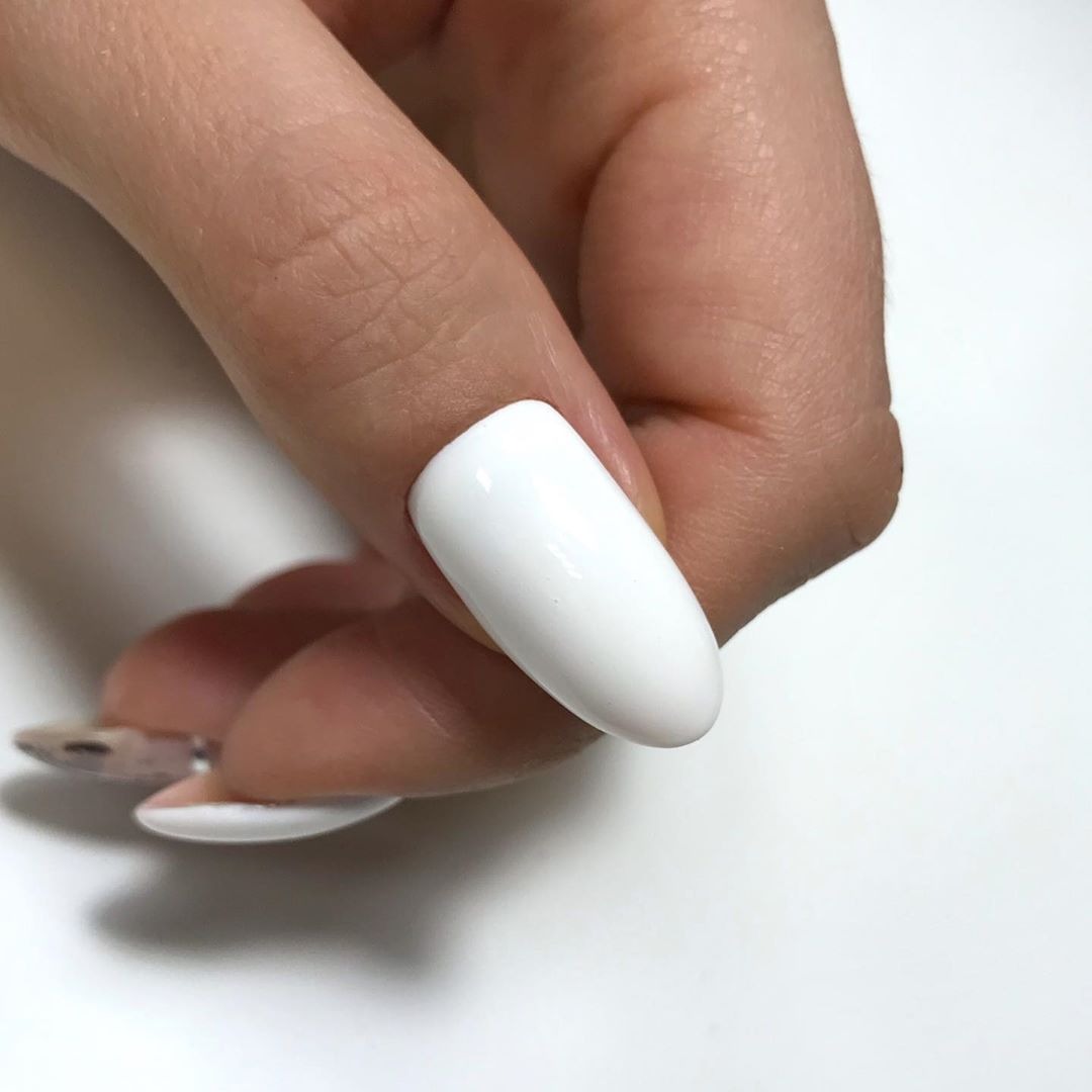 Powiększenie dłoni z jednym pomalowanym na biało paznokciem o owalnym kształcie, pozostałe paznokcie częściowo widoczne, na jasnym tle.