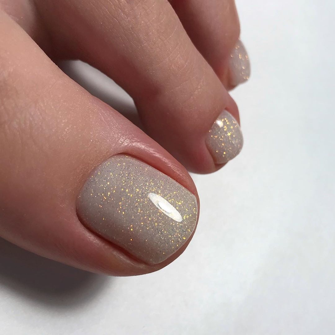 Powiększone ujęcie pomalowanych paznokci u stóp w jasnym, beżowym kolorze z połyskującymi drobinkami, na białym tle.