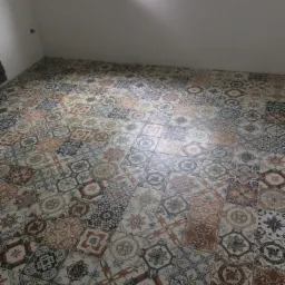 Ułożona podłoga z wzorzystych płytek ceramicznych w odcieniach brązu, beżu i szarości w pustym pomieszczeniu z oknem, na parapecie widoczne akcesoria budowlane.