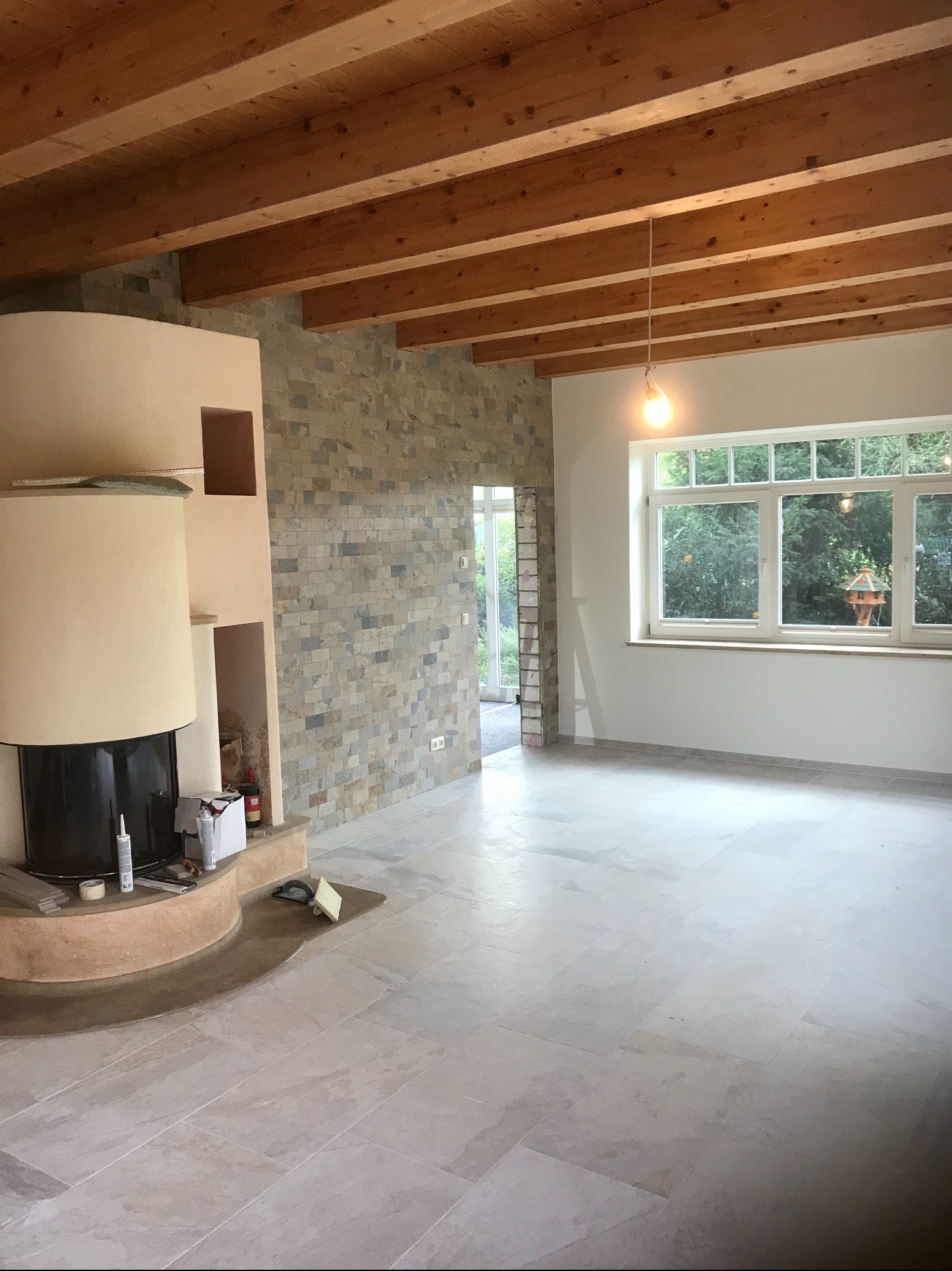 Wnętrze w trakcie remontu: kamienna ściana, sufit z drewnianych belek, kominek z obudową w jasnym kolorze, ułożona podłoga z płytek, widoczne narzędzia i materiały budowlane.